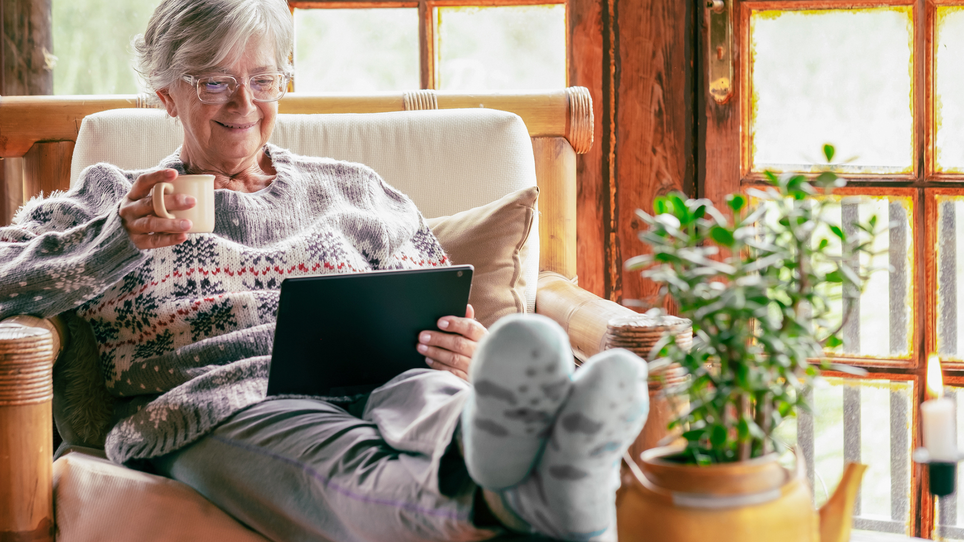 Old senior woman sitting at home on armchair using digital tablet wearing a warm sweater and eyeglasses. Comfortable living room, wooden rustic windows senior woman sitting at home on armchair using digital tablet wearing a warm sweater and eyeglasses